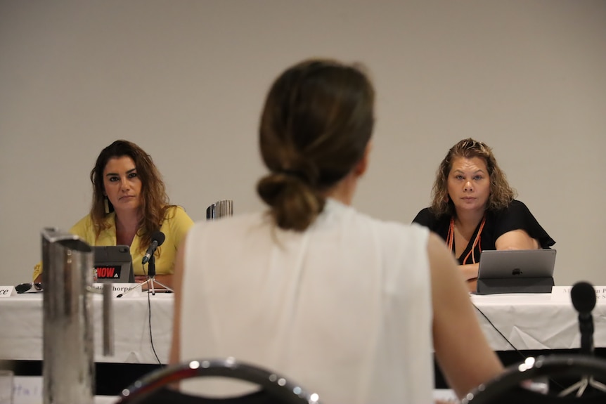 Two women seated at a long table with microphones look seriously ahead of them. 